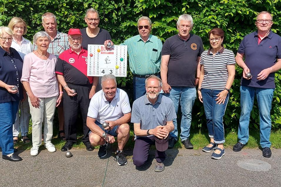 Gruppe von elf Personen posiert lächelnd vor einer grünen Hecke, einige halten Plakate oder Becher in der Hand.