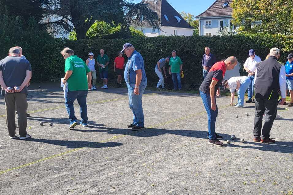 Mehrere Personen spielen Boule auf einem Platz, umgeben von Zuschauern und Bäumen im Hintergrund.