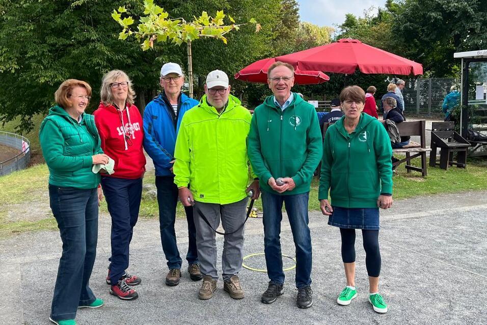 Sechs Personen in grünen Jacken stehen auf einem Platz mit Bäumen und einem Kaffeestand im Hintergrund.