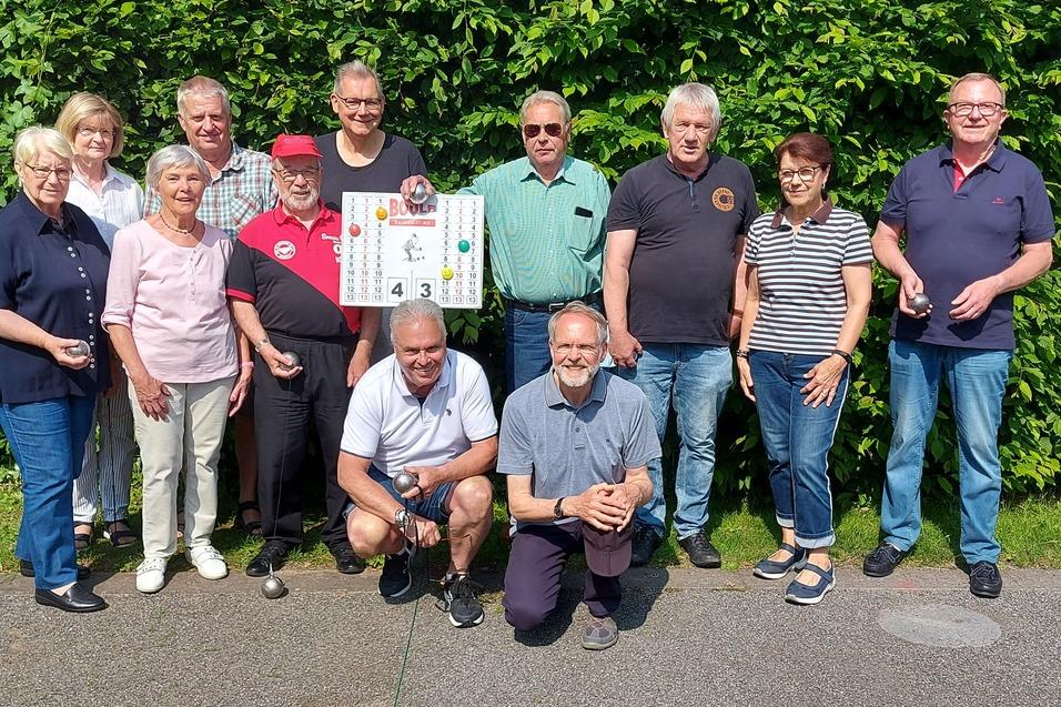Gruppe von elf Personen posiert lächelnd vor einer grünen Hecke, einige halten Plakate oder Becher in der Hand.