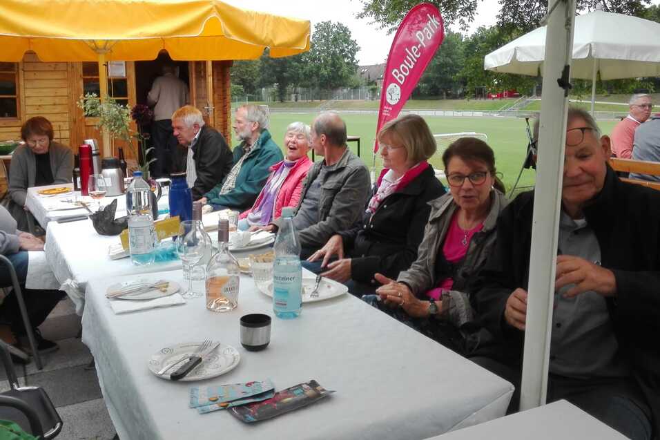 Gruppenszene mit mehreren Personen an einem gedeckten Tisch unter Sonnenschirmen in einem Freiluftbereich.