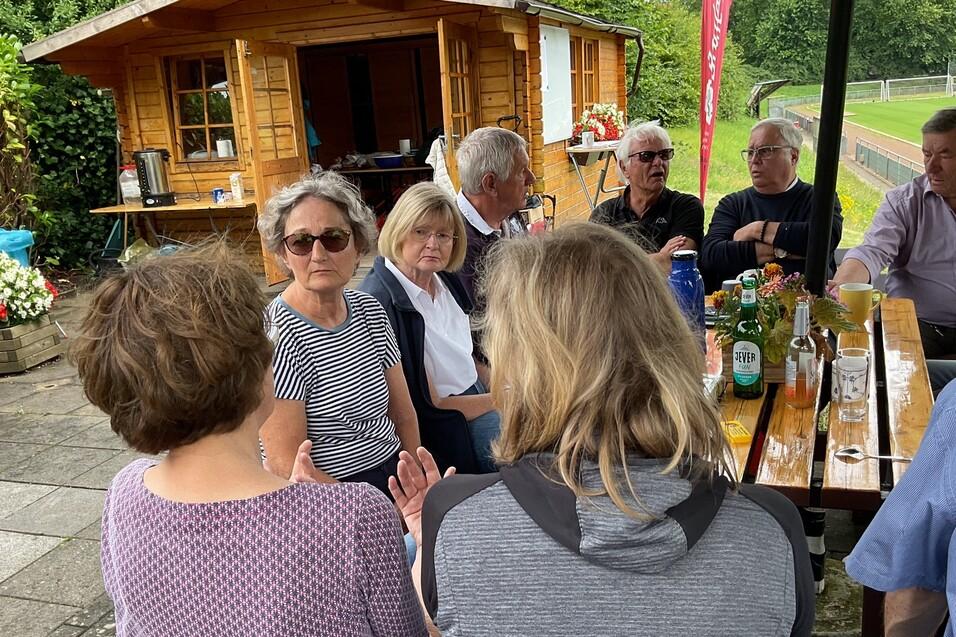 Gruppe von Menschen sitzt an einem Tisch im Freien, spricht und genießt Getränke vor einer Holzhütte.