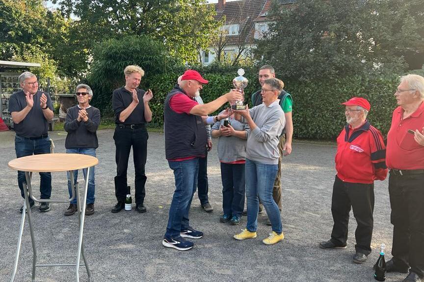 Siegerehrung mit mehreren Personen, die einem Gewinner einen Pokal überreichen, umringt von applaudierenden Zuschauern.