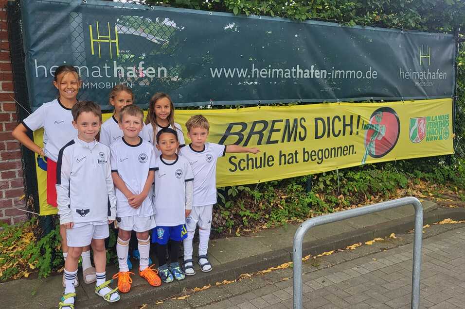Gruppe von Kindern in Fußballtrikots steht lächelnd vor einem Banner mit der Aufschrift „BREMS DICH! Schule hat begonnen.“