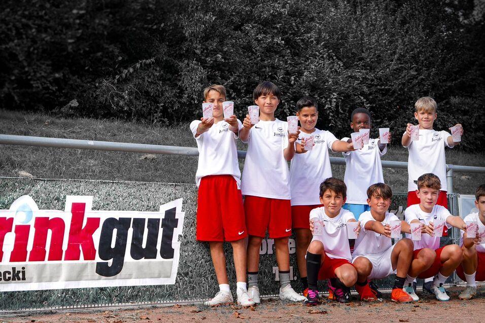 Gruppe von sieben Kindern in Sportkleidung, die stolz im Vordergrund stehen und Zettel in die Kamerarichtung halten, vor Banner.
