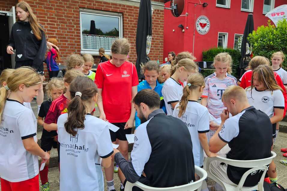 Gruppe von Mädchen in Fußballtrikots umringt Trainer, der Autogramme auf einem Platz vor einem Gebäude gibt.