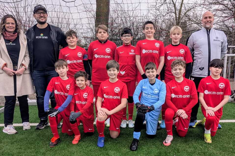 Gruppensfoto einer Jugendfußballmannschaft in roten Trikots mit Betreuern auf einem Sportplatz.