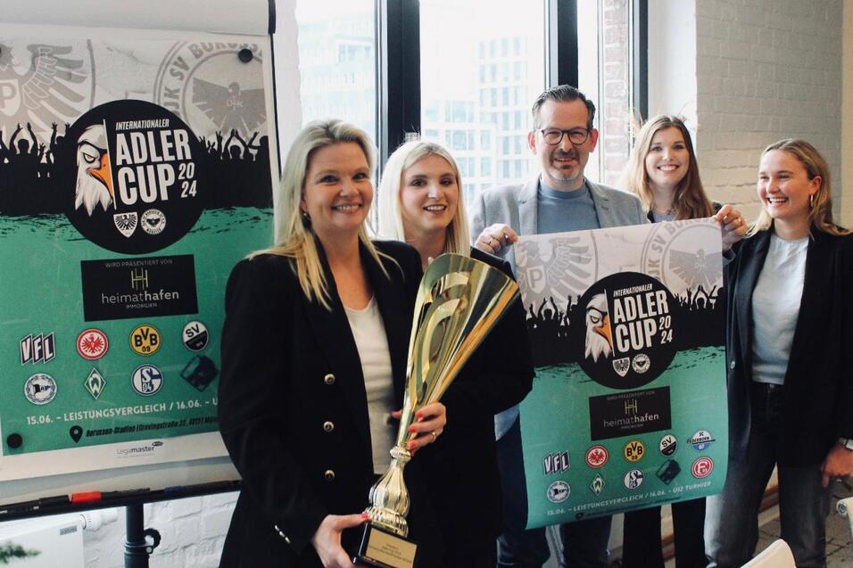 Gruppenszenen mit f&uuml;nf Personen, die mit einer Pokal-Troph&auml;e und einem Banner des Adler Cups 2024 posieren.