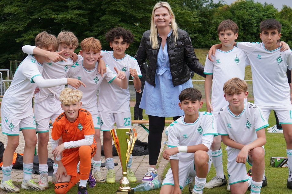 Eine Fu&szlig;ballmannschaft von Jungen posiert mit einem Pokal und einer Trainerin auf dem Spielfeld.