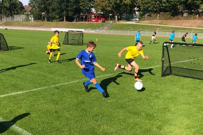 Zwei Kinder in Fußballtrikots rennen auf einem Rasenplatz auf einen Ball zu, mit Toren im Hintergrund.
