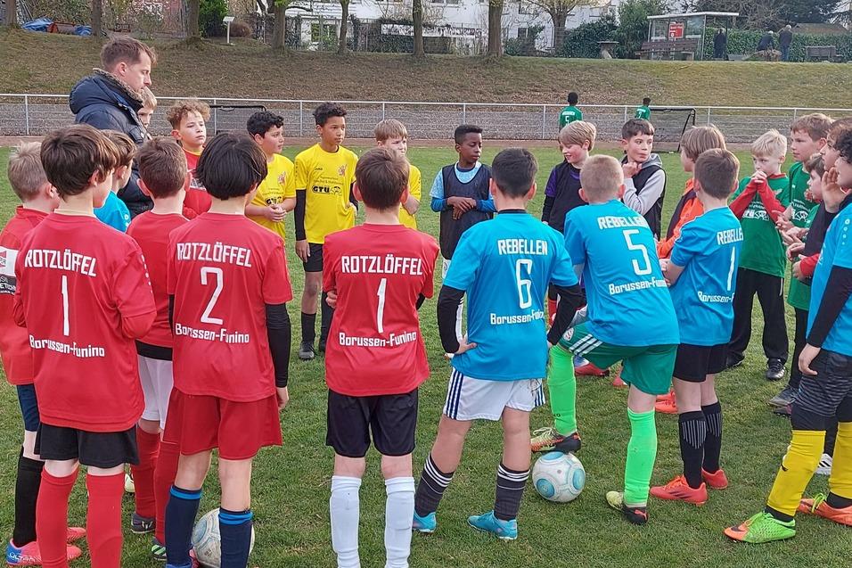 Gruppe von Jugendlichen in Fußballtrikots versammelt sich um einen Trainer auf einem Rasenplatz.