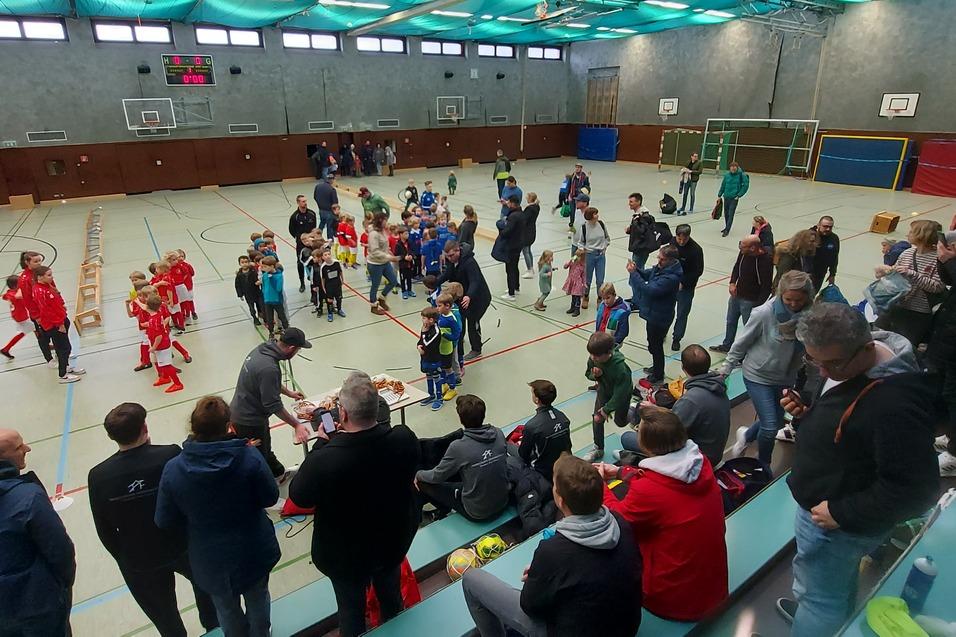 Ein lebhaftes Hallenfußballspiel mit Kindern, Zuschauern und Trainern in einer Sporthalle.
