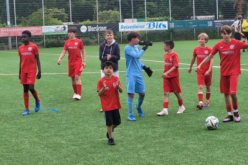 Gruppe von Kindern in roten Fu&szlig;balltrikots auf einem Kunstrasenplatz, einige stehen, andere laufen, ein Torwart in blauer Ausr&uuml;stung.