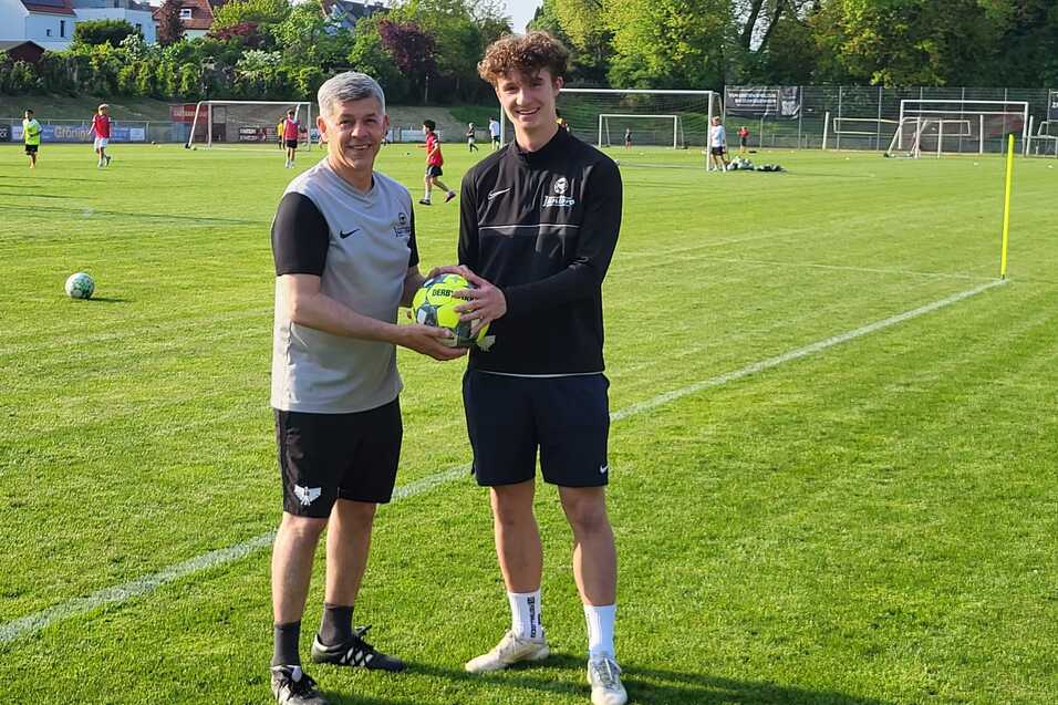 Zwei junge M&auml;nner stehen auf einem Fu&szlig;ballplatz und halten gemeinsam einen Fu&szlig;ball in den H&auml;nden.