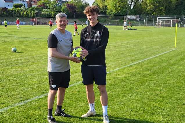 Zwei junge M&auml;nner stehen auf einem Fu&szlig;ballplatz und halten gemeinsam einen Fu&szlig;ball in den H&auml;nden.
