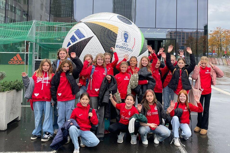 Gruppe von Mädchen in roten T-Shirts posiert vor einem großen Fußball und dem Fußballmuseum, lächelnd und winkend.
