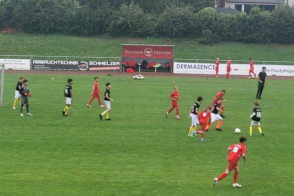 Jugendfußballspiel auf grünem Rasen mit Spielern in roten und schwarzen Trikots sowie einem Tor im Hintergrund.