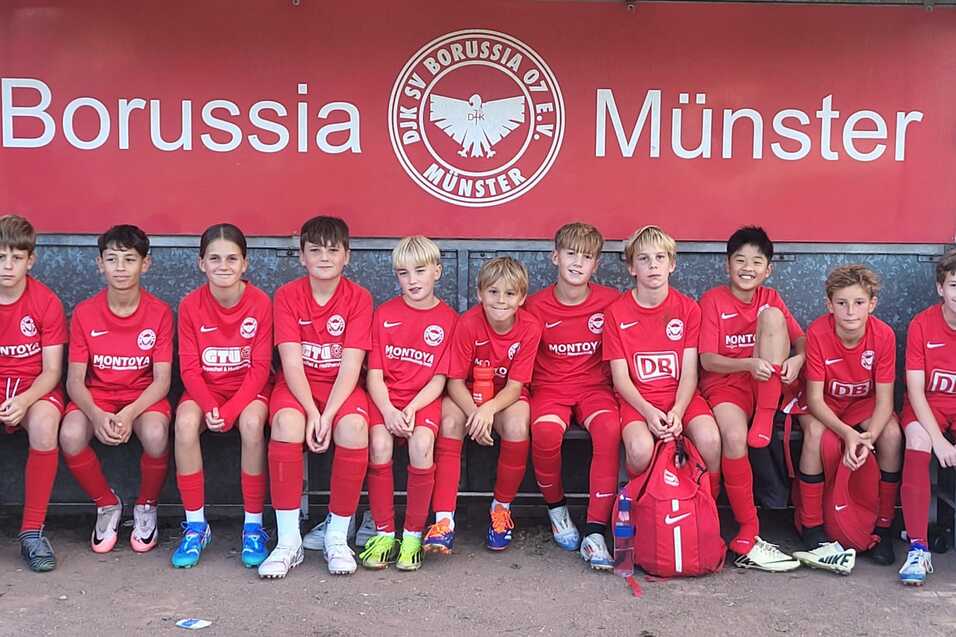 Zehn Kinder in roten Fußballtrikots sitzen auf einer Bank unter einem Borussia Münster-Schild.