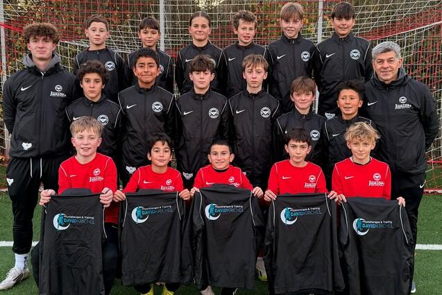 Gruppenfoto einer Jugendfu&szlig;ballmannschaft in sportlicher Kleidung, mit B&auml;llen und Trikots auf einem Fu&szlig;ballplatz.