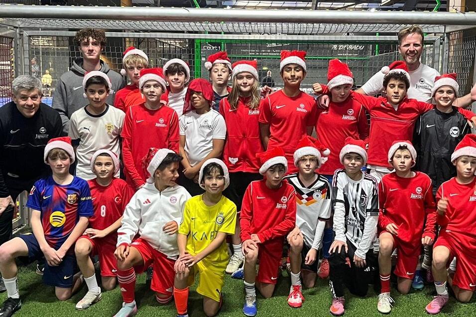 Gruppierung von 20 Kindern in Fu&szlig;balltrikots und Weihnachtsm&uuml;tzen vor einem Tor auf einem Sportplatz.