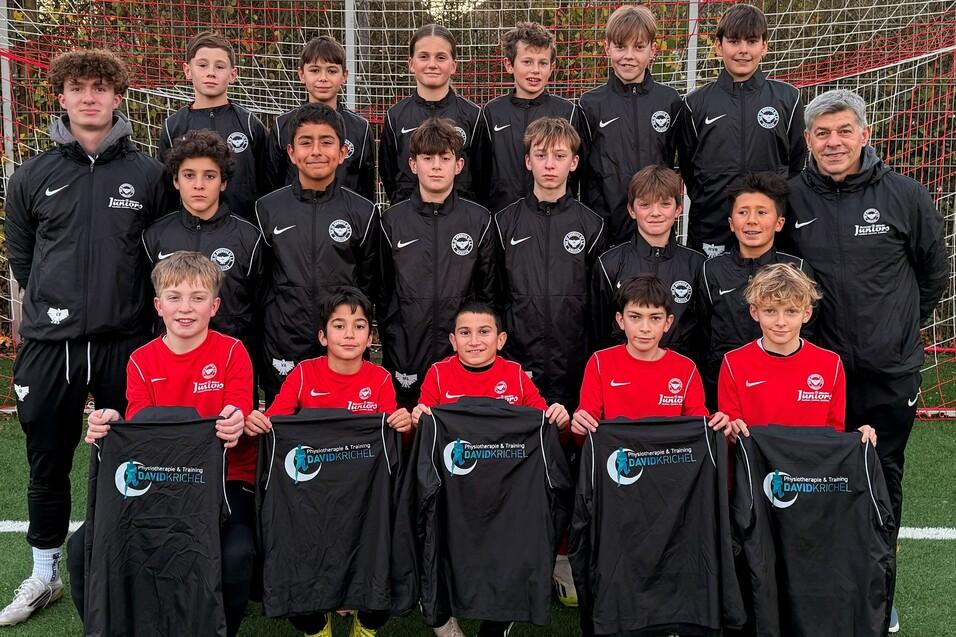 Gruppenfoto einer Jugendfu&szlig;ballmannschaft in sportlicher Kleidung, mit B&auml;llen und Trikots auf einem Fu&szlig;ballplatz.