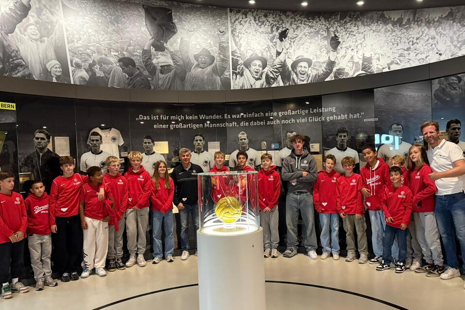 Gruppe von Kindern in roten Jacken steht um eine Glaskuppel mit einem Ball, im Hintergrund sportliche historische Bilder.