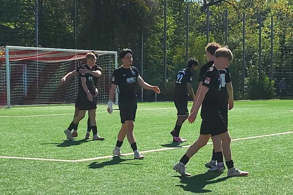 Fußballspiel auf Rasenplatz mit Spielern in schwarzen und roten Trikots, Tornetz im Hintergrund.