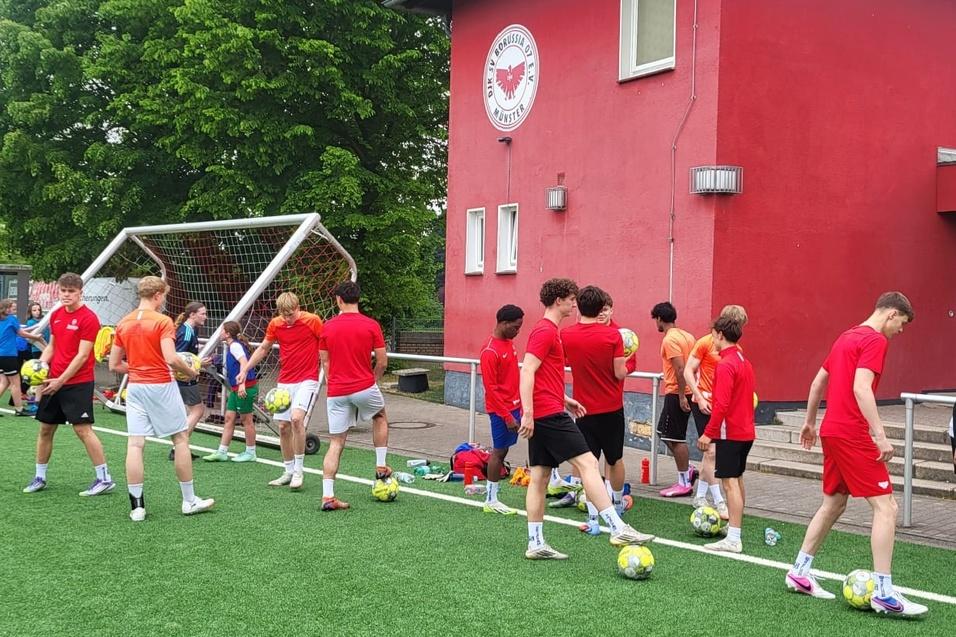Gruppe junger Sportler in roten T-Shirts trainiert auf einem Fußballplatz, umgeben von Bällen und Toren.