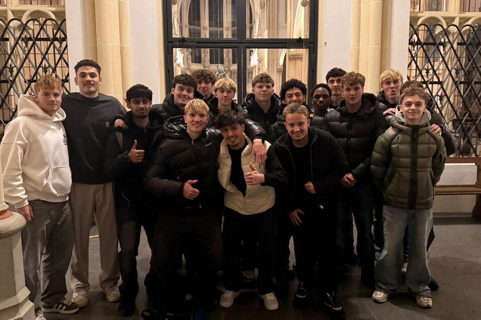 Gruppe von Jugendlichen in sportlicher Kleidung posiert lächelnd in einem historischen Innenraum mit großen Fenstern.