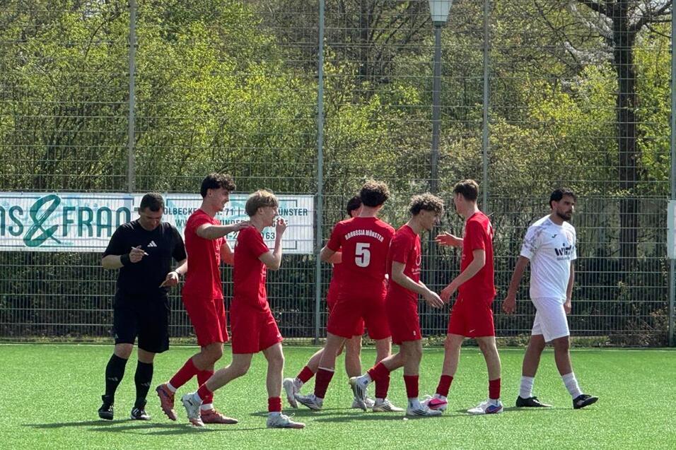 Sechs Fußballspieler in roten Trikots stehen auf einem Kunstrasenplatz, während ein Schiedsrichter in Schwarz daneben steht.