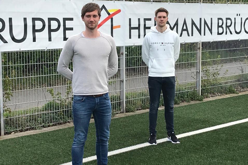 Zwei junge Männer stehen auf einem Kunstrasenfeld vor einem Banner mit der Aufschrift „GRUPPE HERMANN BRÜCK“.