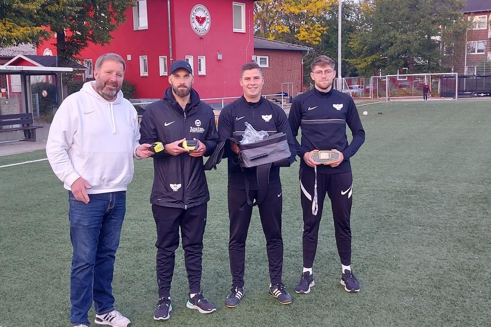 Vier Personen stehen auf einem Fußballplatz; zwei Spieler halten Preise, während ein Trainer daneben steht.