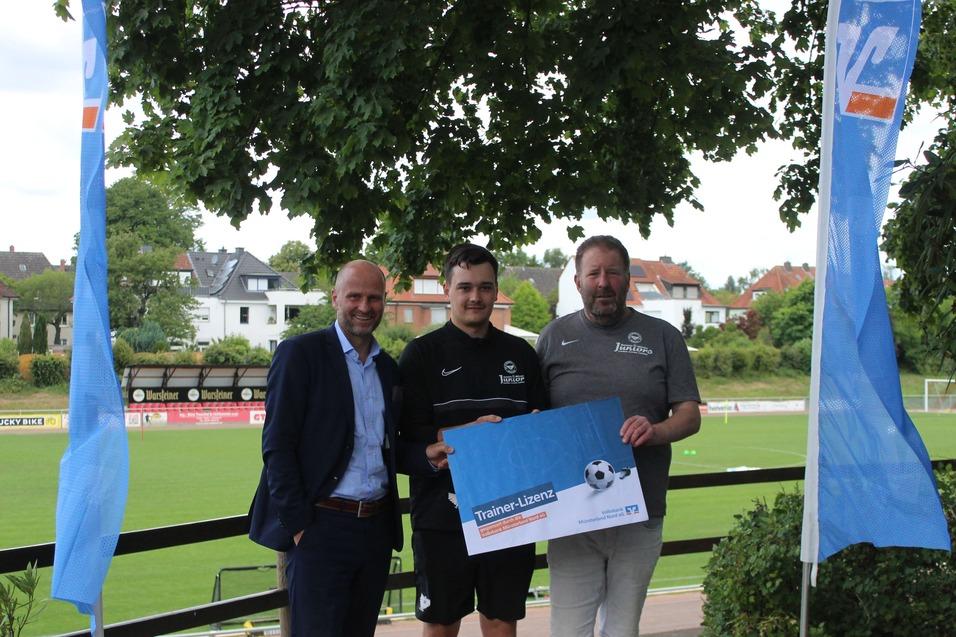 Drei Männer stehen auf einem Sportgelände, halten ein Plakat und posieren vor einem Fußballplatz mit Bäumen im Hintergrund.