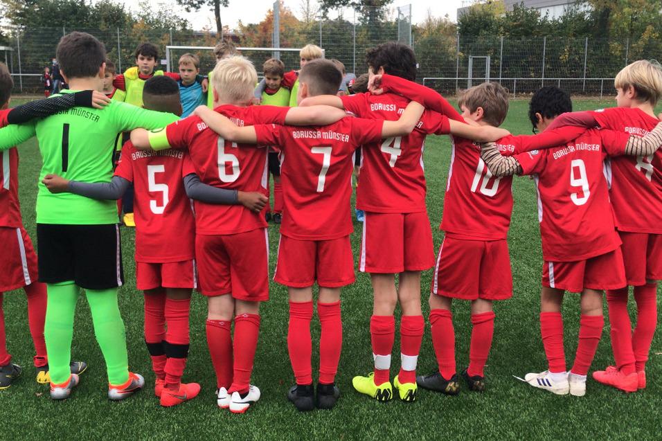 Eine Gruppe junger Fu&szlig;ballspieler in roten Trikots steht mit dem R&uuml;cken zueinander und umarmt sich auf einem Kunstrasenplatz.