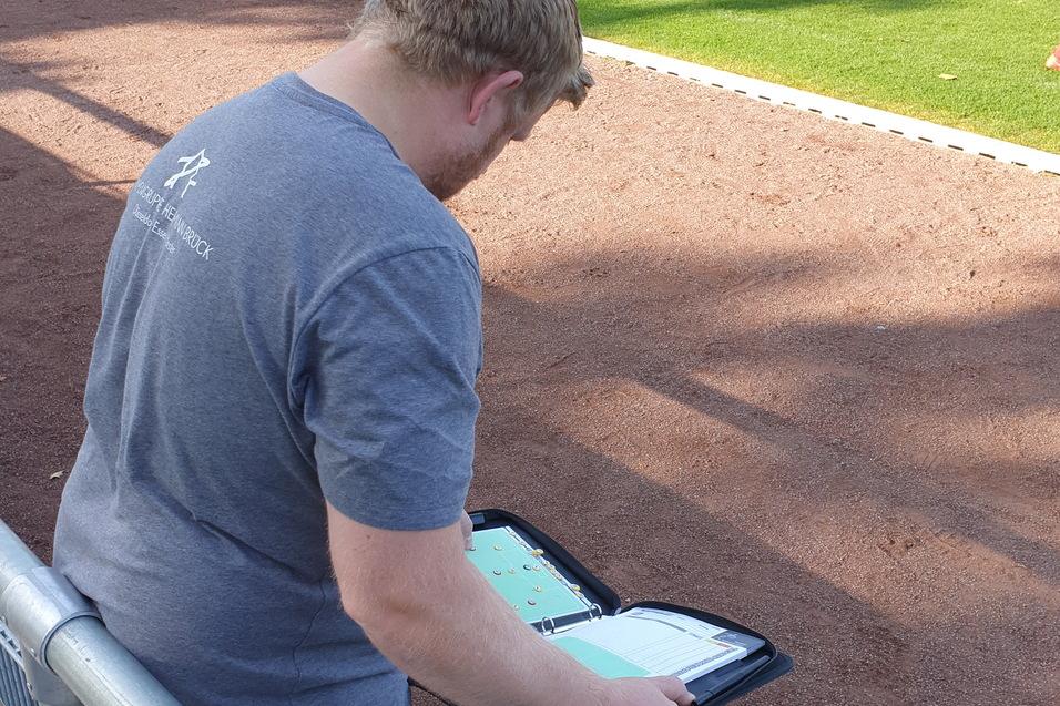Ein Trainer beobachtet ein Fu&szlig;ballspiel und notiert auf einem Tablet, w&auml;hrend Spieler auf dem Feld trainieren.
