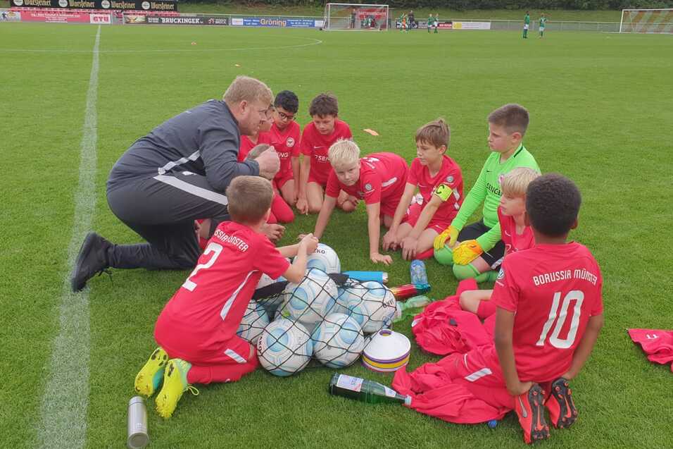 Gruppe von Kindern in roten Trikots sitzt um B&auml;lle und Ausr&uuml;stung auf einem Fu&szlig;ballfeld, w&auml;hrend ein Trainer spricht.