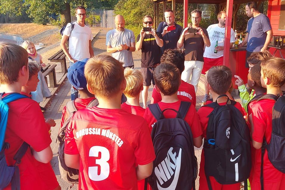 Gruppe von Kindern im roten Fu&szlig;balltrikot h&ouml;rt einem Trainer an einem Vereinsheim zu.