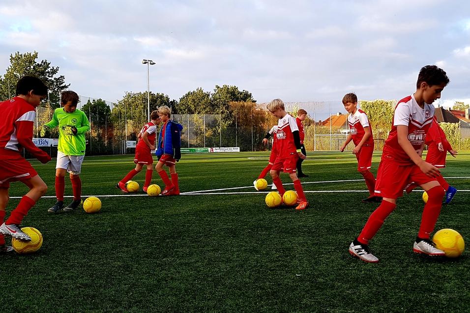 Jugendliche Fußballspieler in roten Trikots üben mit gelben Bällen auf einem Kunstrasenplatz unter bewölktem Himmel.