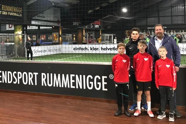 Gruppe von vier Jungs in roten Sportjacken posiert mit einem Erwachsenen in einer Soccer-Halle vor einem Fu&szlig;ballfeld.