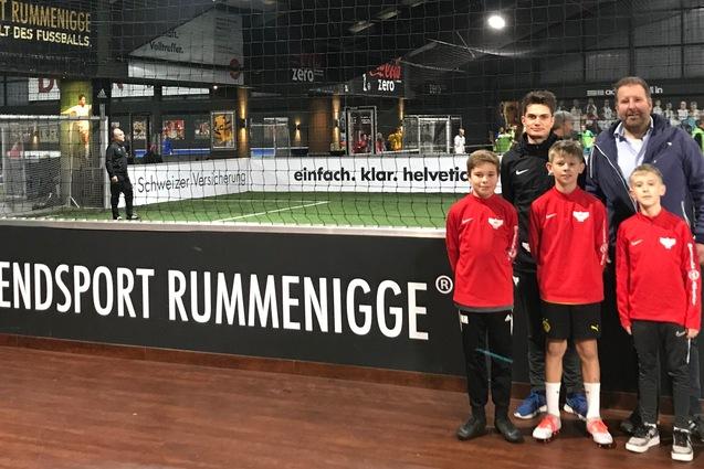 Gruppe von vier Jungs in roten Sportjacken posiert mit einem Erwachsenen in einer Soccer-Halle vor einem Fu&szlig;ballfeld.