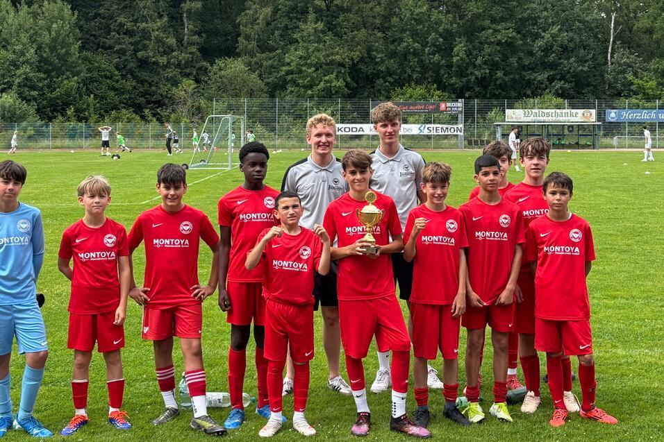 Gruppe von elf Spielern in roten Trikots und einem Trainer auf einem Fu&szlig;ballplatz, halten einen Pokal in den H&auml;nden.