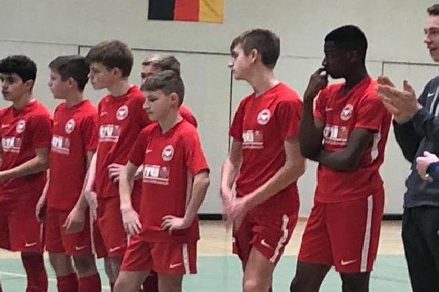 Eine Gruppe von sieben Jugendlichen in roten Fu&szlig;balltrikots steht an der Seitenlinie eines Hallenfu&szlig;ballspiels.