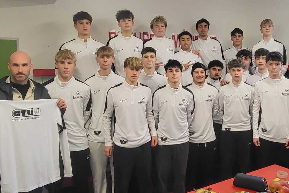 Gruppe von 16 jungen Sportlern in Trainingsanzügen, die in einem Umkleideraum mit einem Trainer posieren.
