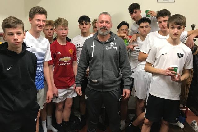 Gruppe von jungen Fu&szlig;ballspielern in einem Umkleideraum, umrahmt von einem Trainer in sportlicher Bekleidung.