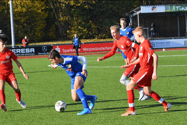 Vier jugendliche Fu&szlig;ballspieler im Wettkampf auf einem Kunstrasenplatz, einer dribbelt den Ball, w&auml;hrend die anderen versuchen zu verteidigen.