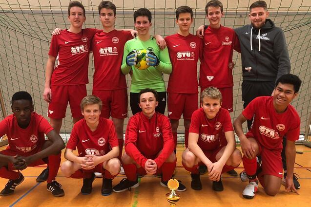 Jugendfu&szlig;ballmannschaft in roten Trikots posiert mit Pokal vor einem Tor in einer Sporthalle.