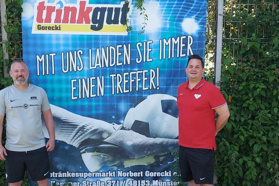 Zwei Männer stehen vor einem Werbebanner mit Fußball-Motiv und dem Text „Mit uns landen Sie immer einen Treffer!“