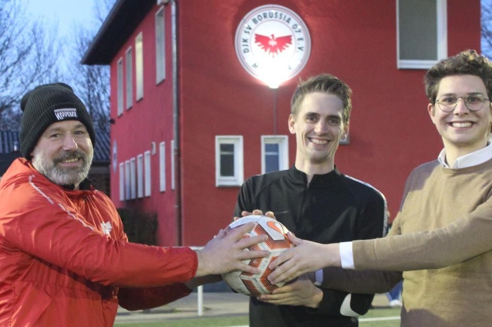 Drei Männer stehen auf einem Fußballplatz und übergeben gemeinsam einen Fußball vor einem roten Vereinsgebäude.