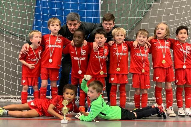 Eine Gruppenaufnahme von zw&ouml;lf jubelnden Kindern in roten Fu&szlig;balltrikots und zwei Trainern vor einem Tor, mit Medaillen.