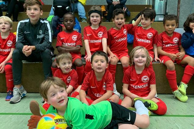 Gruppe von Kindern in roten Fu&szlig;balltrikots, sitzend auf einer Trib&uuml;ne, l&auml;cheln in die Kamera. Ein Junge tr&auml;gt ein Torwart-Trikot.